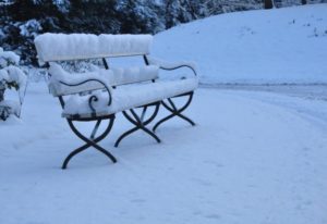 A metal park bench covered in a thick layer of snow, surrounded by untouched snow in a quiet outdoor setting.