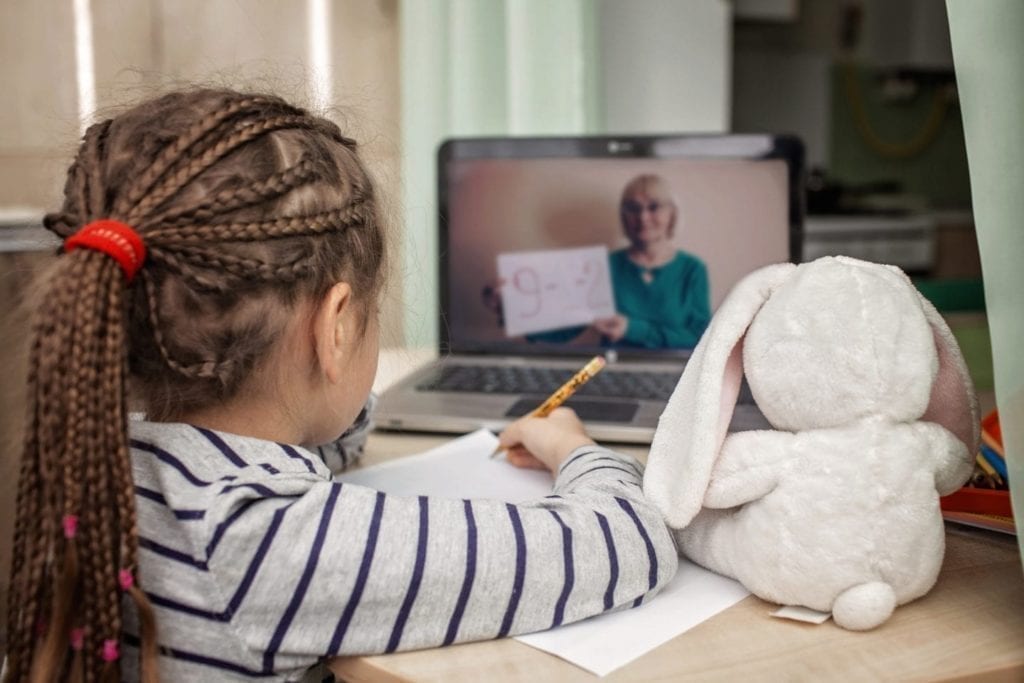 A child writes on paper at a desk, her stuffed bunny beside her, as she watches a woman holding a math problem on a laptop screen during a Back to School online lesson.