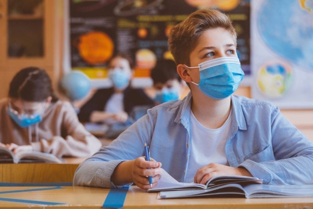 Back to School: Students wearing face masks sit at desks in a classroom, writing in notebooks and appearing attentive, with educational posters visible in the background.