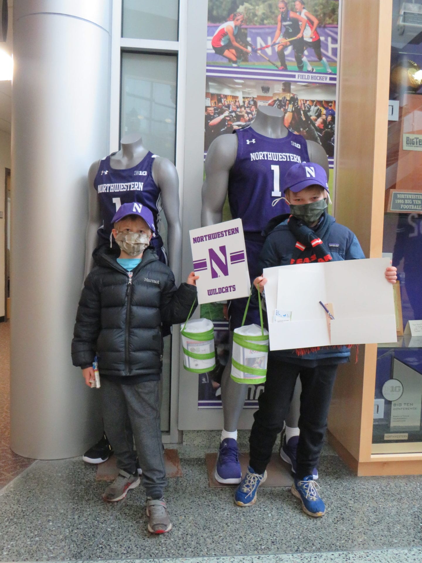Two children wearing masks stand next to a Northwestern Wildcats mannequin display; one holds a northwestern football team sign, the other holds a blank poster and buckets.