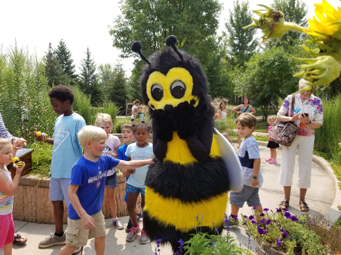 A person in a bee costume stands on a path surrounded by smiling children and adults, while the Bug Girls lead garden activities in an outdoor setting on a sunny day.