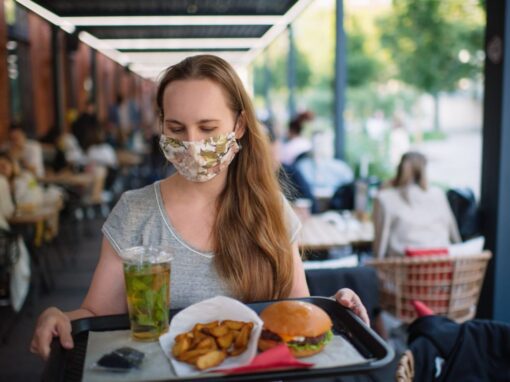 A woman wearing a floral face mask carries a tray with a burger, fries, a drink, and sauce at an outdoor restaurant—a glimpse into the Food Industry’s adaptations.