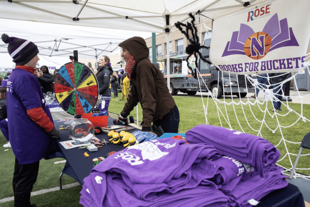 A child spins a prize wheel at an outdoor booth with purple t-shirts and books, under a banner announcing "Books for Buckets is Back." Adults staff the booth.