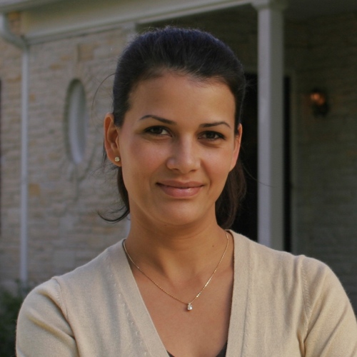 Woman with dark hair pulled back, wearing a light sweater and necklace, standing outside in front of a stone building with white trim.