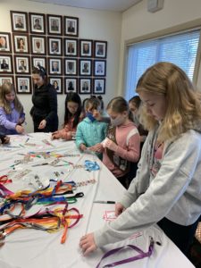 A group of children and an adult gather around a table with colorful lanyards and name tags, working on crafts during a Girl Scout Spark Day in a room decorated with framed photos on the wall.