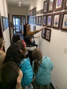 A woman points to a framed photo on a wall of portraits while a group of children in jackets gather around her in the hallway during Girl Scout Spark Day.