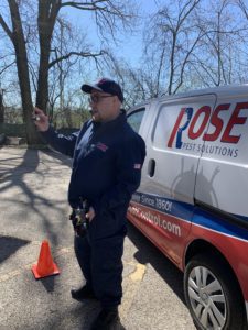 A pest control worker in uniform stands next to a Rose Pest Solutions van, holding an object and speaking outdoors near a traffic cone during Girl Scout Spark Day.