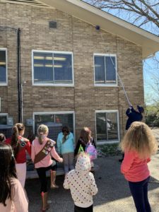 During a Girl Scout Spark Day, a person uses a long pole to retrieve an object from the roof of a brick building while a group of children watches from below.