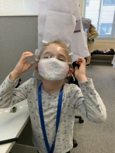 Young child, possibly at Girl Scout Spark Day, wearing a sparkly face mask and clear protective glasses, stands indoors by a desk, looking up with hands at their face.