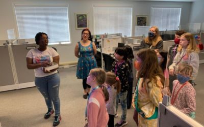 A group of children and adults, some wearing masks, listen to a woman speaking in an office setting with cubicles and windows during a Girl Scout Spark Day.