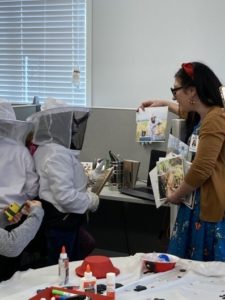 Two children in beekeeping suits stand beside a woman holding up a photo; art supplies are spread out on a nearby table during Girl Scout Spark Day.