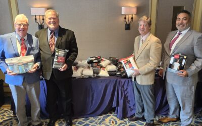 Four men in suits stand indoors, each holding packages of socks and shirts, in front of a table piled with similar clothing items.