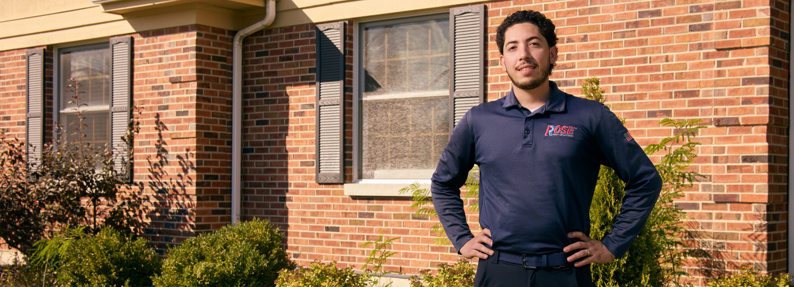 251008_rose-E_0004_w A man in a navy blue PASE polo shirt stands outside a brick house with his hands on his hips