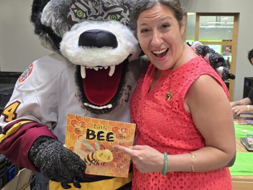 A woman in a red dress stands next to a wolf mascot, holding a children's book titled