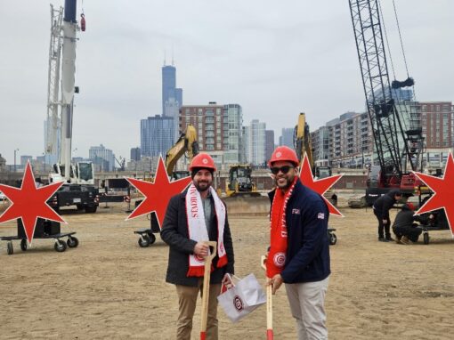 Wesley and Andrew in hard hats and scarves stand with shovels at a Chicago Fire construction groundbreaking site, with city buildings and construction equipment in the background.