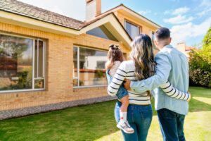 A couple stands outside holding a young child, looking at a modern brick house on a sunny day.