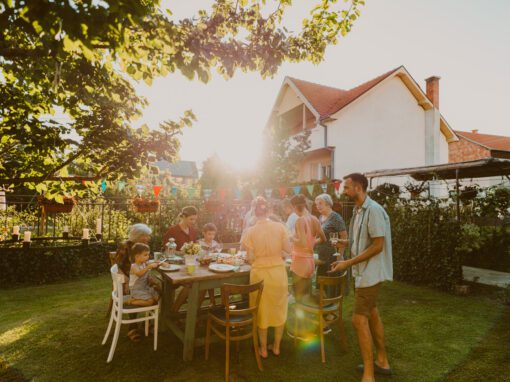 A group of people gather around a table for an outdoor meal in a backyard, with sunlight streaming through trees, colorful bunting overhead, and the occasional buzz of flies in summer.
