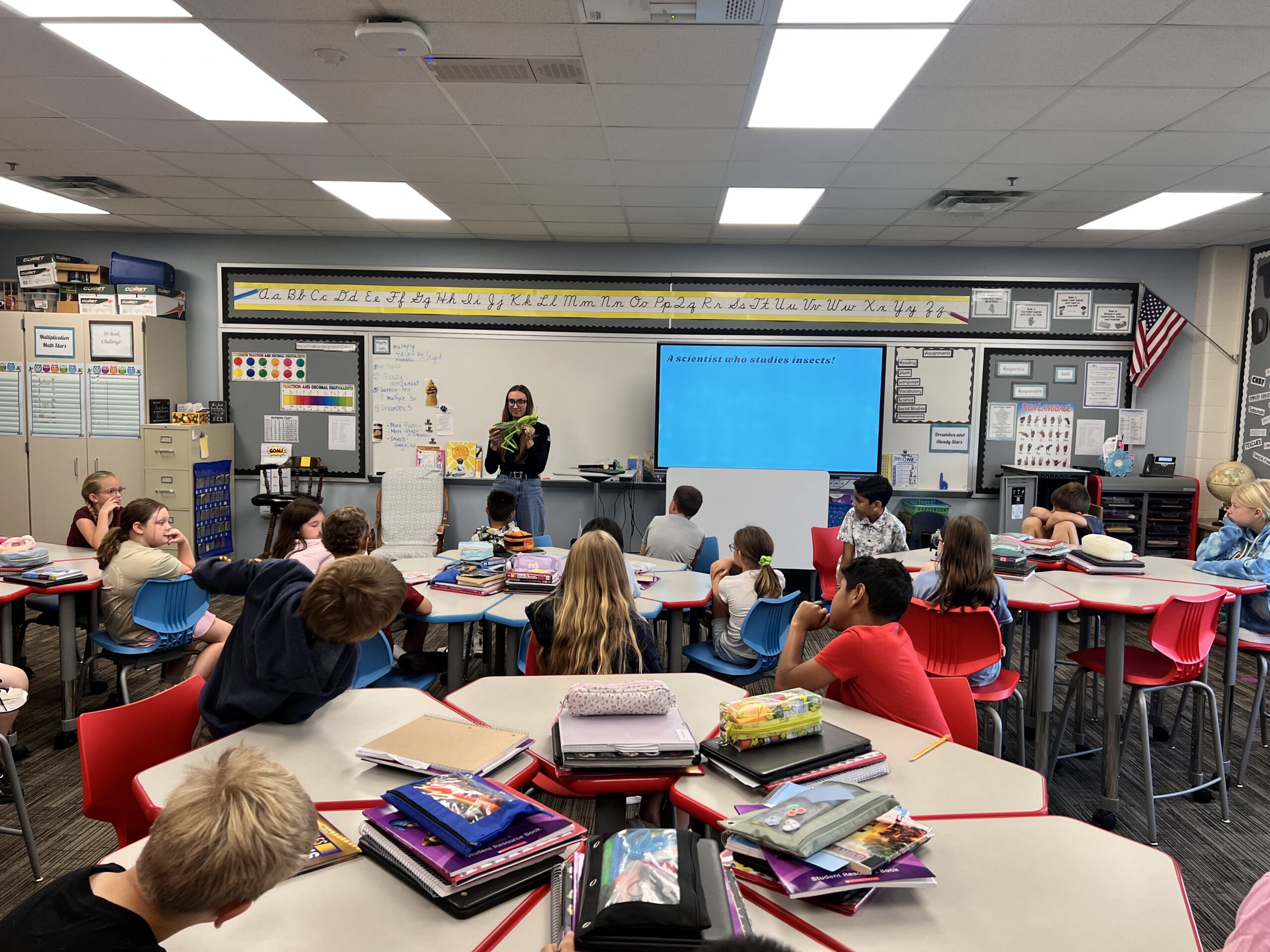 Allison stands at the front of a classroom with students seated at round tables, facing a screen displaying a blue slide featuring “The Bug Squad.” Classroom supplies and notebooks are visible on the tables.